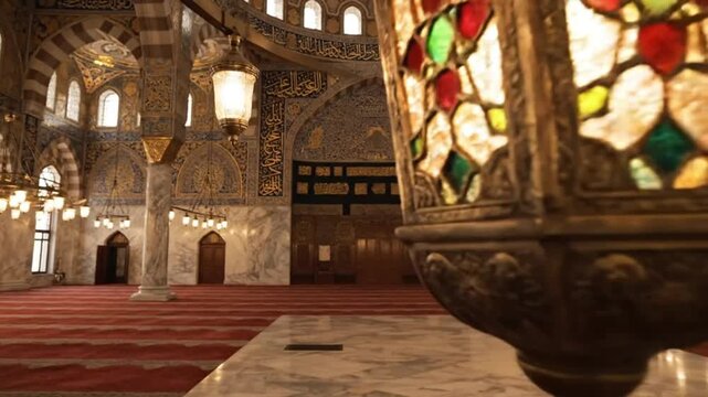 Ornate lamp in the foreground with stained glass, illuminating the interior of a grand mosque with red carpet and intricate architectural details