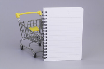Shopping cart with blank notebook page on grey background. List of goods to buy concept.