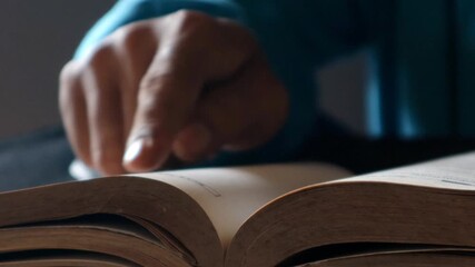 Close-up of male hands turning book page, nestled atop stacked volumes in soft, muted library setting, creating intimate reading environment, selective focus.