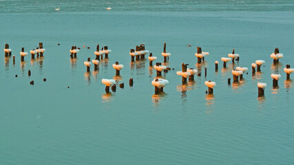 Wooden posts covered with ice and snow standing in calm turquoise water during winter. Minimalist cold landscape with reflections, frozen lake surface and natural patterns. Concept of winter calm