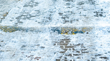 Icy brick pavement with snow, slush and a yellow-black speed bump partially covered by ice. Winter urban background showing slippery road conditions, frozen surface and pedestrian hazard. 