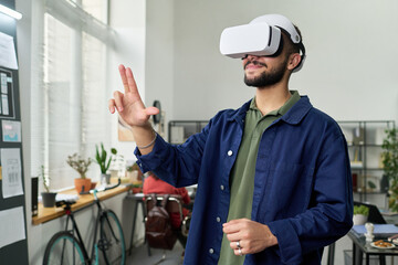Young adult man wearing virtual reality headset interacting with digital interface in modern office setting, standing near window and using hand gestures for navigation