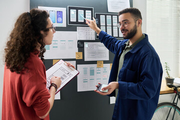 Young adult man explaining mobile app interface designs to male colleague holding clipboard, both standing in front of wall with wireframe sketches and diagrams