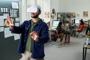 Young adult man using virtual reality headset and interacting with digital interface in modern office while diverse coworkers working at desks in background
