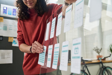 Young adult Caucasian man arranging mobile app interface mockups on transparent board, standing in modern office, focusing on user experience design process for digital project