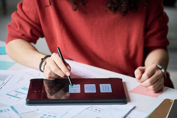 Midsection of man using digital pen to design mobile app interface on tablet, working at desk with paper wireframes and mockups, focusing on creative process in modern workspace