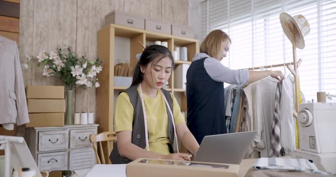 slowmotion scene asian female dressmaker working on laptop fashion studio while team member checking clothing rack behind sewing tools packaging boxes arranged on wooden shelves startup business