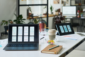 Modern office workspace showing open laptop and tablet displaying mobile app interface prototypes, takeaway coffee cup, notebook, documents, and bowl of colorful candies on desk