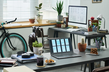 Modern workspace featuring open laptop displaying wireframe designs on desk, desktop monitor with interface layouts in background, bicycle near window, various plants and office supplies arranged