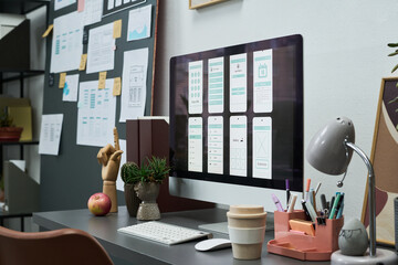 Modern workspace showing computer monitor displaying mobile app interface designs, desk with office supplies, coffee cup, and plants, wall covered with wireframes and sketches in background
