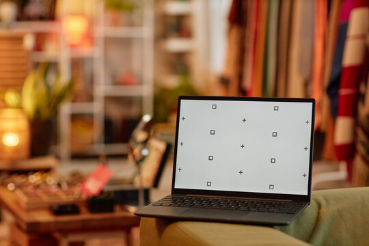 Open laptop displaying calibration screen standing on sofa in second hand shop, blurred racks of used clothing and accessories visible in background, no people present