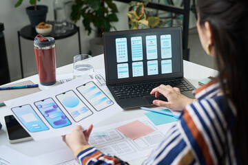 Young adult woman designing mobile app interface, holding wireframe sketches while working on laptop displaying smartphone screen layouts, sitting at desk with digital tablet and documents © pressmaster