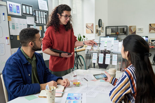 Multiethnic group of young adults collaborating on mobile app interface design, discussing wireframes and digital prototypes at creative office workspace, using tablet and printed layouts - Powered by Adobe