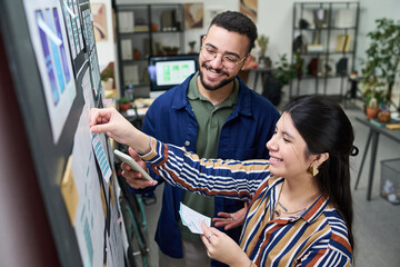 Young man and woman collaborating on creative project, smiling while organizing design mockups on wall, holding smartphone and paper materials