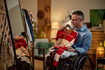 Middle aged Caucasian man with disability sitting in wheelchair holding second hand striped sweater, inspecting garment in front of mirror in cozy indoor setting surrounded by clothing