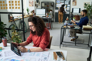 Caucasian young adult man using digital tablet while working on design project at desk in modern office, multiethnic young adult colleagues collaborating in background