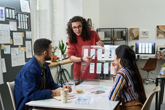 Young adult Caucasian man presenting mobile app wireframes to young adult multiethnic colleagues in modern office, team collaborating on user interface design project, discussing prototypes - Powered by Adobe