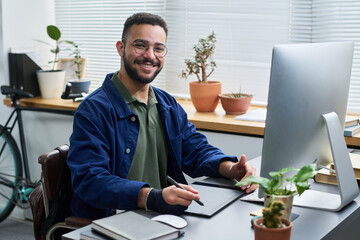 Portrait of young adult man with beard smiling at camera, while sitting at desk using digital drawing tablet in modern office workspace with plants and computer monitor