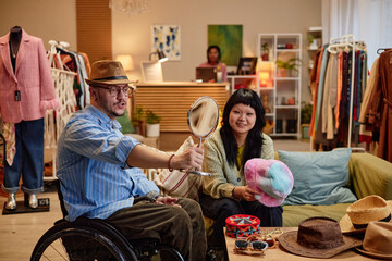 Caucasian middle aged man with disability sitting in wheelchair trying on hat in second hand shop, while Asian young adult woman holding colorful hat sitting beside him smiling