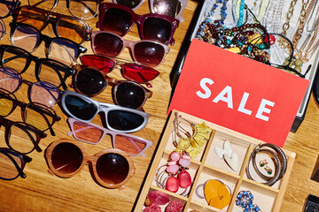 Assorted second hand sunglasses and jewelry displayed on wooden table with red sale sign, various styles and colors arranged for discount shopping at thrift store or flea market