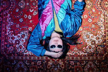 Portrait of young adult Asian woman lying on vintage patterned carpet wearing sunglasses, holding head with one hand, embodying retro style and second hand fashion concept