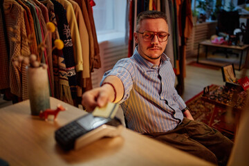 Obraz premium Caucasian middle aged man sitting at counter making payment with credit card in second hand clothing store, shelves with vintage garments and accessories visible in background