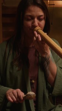 Medium vertical of inspired Caucasian female esoteric music performer sitting with crossed legs in dimly candle lit yoga studio, playing didgeridoo and rattle during sound meditation practice