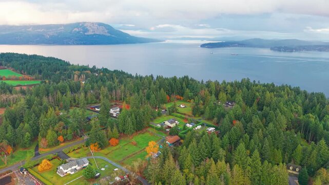 Aerial drone footage of forested residential homes overlooking the calm waters of Saanich Inlet near Mill Bay, British Columbia, with mountains and islands in the background during autumn.