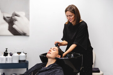 Professional hairstylist washing hair of woman in modern beauty salon, soothing cleansing treatment in white interior