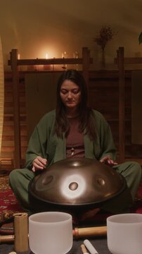 Full vertical shot of young Caucasian female musician playing music on handpan, while performing at private candlelit concert, singing bowls, wind chime, didgeridoo lying on carpet
