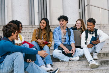 Obraz premium Group of diverse university students smiling and chatting happily while sitting on campus stairs outside the building