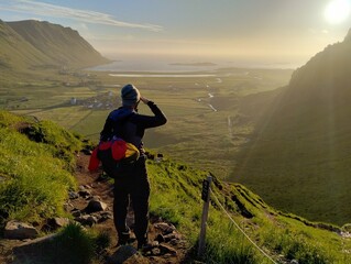 Hiker with backpack standing on a mountain trail and looking over a green valley in golden hour...