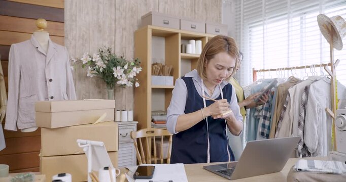 asian female adult fashion designer organizing cardboard boxes inside tailoring studio workspace preparing handmade clothing product shipping packing process under natural daylight near sewing machine