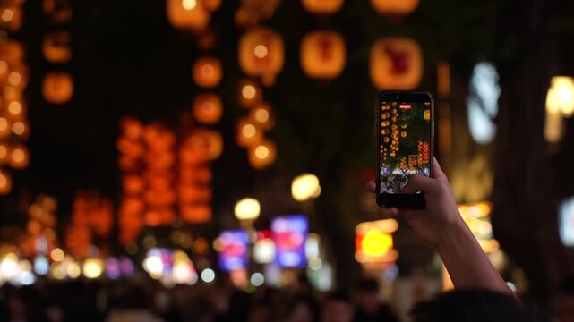 A person recording a lantern-decorated pedestrian street with a smartphone in Guangzhou at night. The phone screen is in focus while warm glowing lanterns and cit