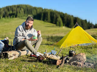 Man tourist eating from food pouch with green spoon, sits on log near campfire. Yellow tent on green hillside, surrounded by lush trees. Peacefulness of camping in beautiful natural environment.