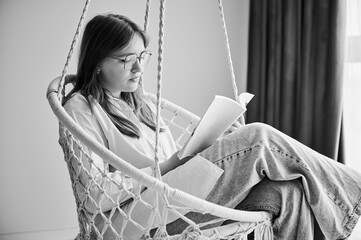 Concentrated girl relaxing and reading book on indoor swing. Pretty female in glasses excited about story she reading. Cozy afternoon with book in hanging chair in home. Black and white image.