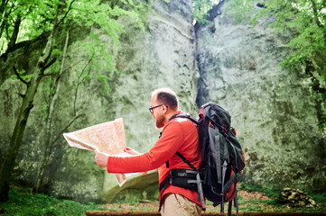 Bearded tourist man studies map while stands in front of large rock in dense forest. Traveler with grey backpack and glasses, planning outdoor adventure, hiking or climbing route.