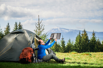 Man sits beside tourist tent, holding portable solar panel charger to capture sunlight for charging power bank. Large orange backpack, camera on tripod. Snow-capped mountains under partly cloudy sky.