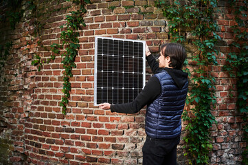 Man holding photovoltaic solar panel in front of old brick wall covered in green ivy. Concept of integration of sustainable renewable energy sources into historical architecture.