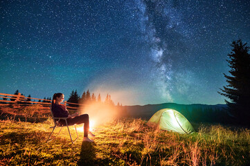 Hiker woman relaxes by campfire, illuminated by its warm glow, with tent nearby under stunning, star-filled sky. Milky Way stretches overhead, adding magical touch to serene mountain setting.