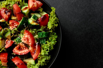 Vegetable salad of tomatoes, cucumbers, onions, lettuce dressed with aromatic dressing in a black plate on a gray background
