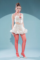 Elegant Asian model in white lace corset top and ruffled mini skirt with red ribbon accents, posing confidently in sheer red stockings and nude high heels, glamorous studio portrait soft blue scene