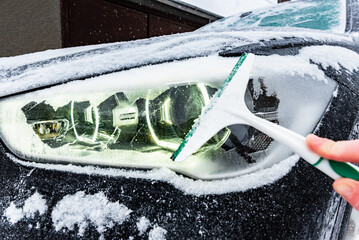 Woman's hand removing snow and scraping ice from car front headlight in winter.