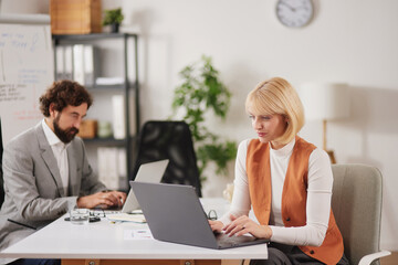 Team members sit at a desk in an office discussing ideas and working on tasks together while using...