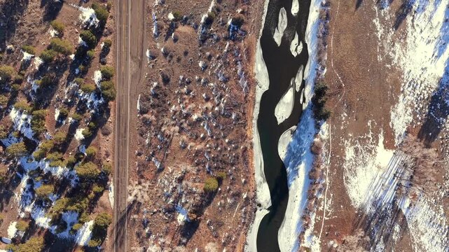 A cinematic top-down aerial shot follows a paved highway running parallel to a partially frozen river. This snowy winter landscape is perfect for automotive commercials and travel brand storytelling.