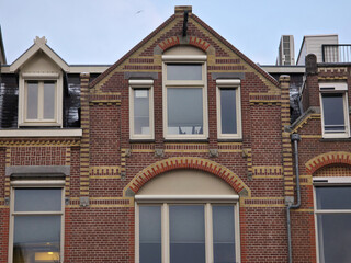 A brick building facade typical of Amsterdam, featuring detailed yellow-striped brickwork, diverse window styles including arched ones, and pitched roofs with dormers and skylights
