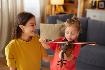 A young girl is learning to play the violin with her mother. They are both smiling and sitting on the floor of their cozy living room, enjoying this special family moment. © Stockphotodirectors