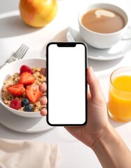 Hand holds a blank phone above a breakfast scene of oatmeal, juice, coffee, and apple in bright, morning light