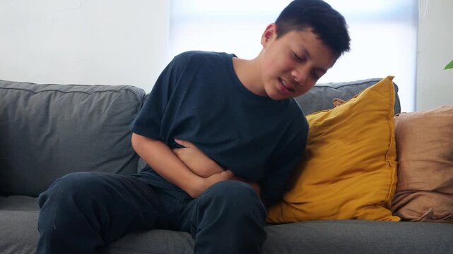 Asian Young Boy Clutching His Abdomen Suffering from Painful Stomach Ache on Sofa