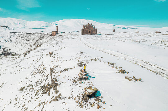 Kars, Ani Ruins: An aerial winter view of the snow-covered medieval ruins along the Arpacay River on the Turkey&ndash;Armenia border. A historical landscape featuring a man contemplating the scene.